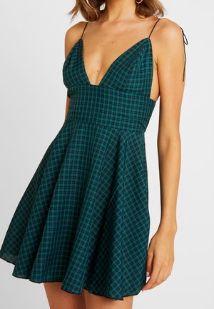 Femme portant une robe sans manches à carreaux verts et noirs, avec un profond décolleté en V et des fines bretelles nouées dans le dos.