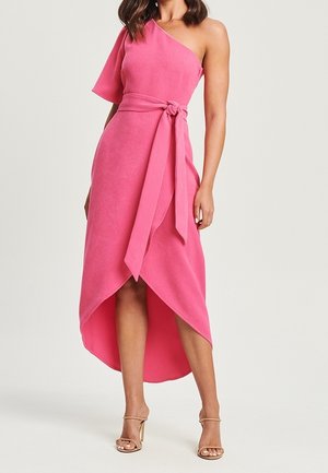 Kobieta w różowej sukience na jedno ramię z wiązanym w pasie i asymetrycznym dołem, połączona z beżowymi sandałami na wysokim obcasie.