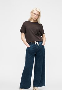 Νέα γυναίκα με ξανθά μαλλιά που φοράει σκούρο καφέ κοντομάνικη μπλούζα, φαρδιά μπλε τζιν με λευκό κορδόνι και αθλητικά παπούτσια, στέκεται με τα χέρια στις τσέπες.