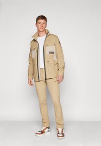 Giacca beige con tasche chiare a contrasto, zip completa e colletto regolabile, abbinata a pantaloni beige coordinati e sneaker multicolori.