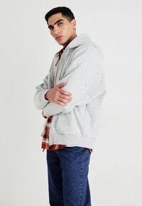 Joven con los brazos cruzados, vistiendo una sudadera gris clara sobre una camisa de cuadros roja y jeans azules, de pie frente a un fondo blanco liso.