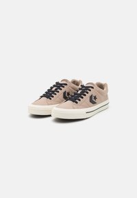 Baskets beiges avec des lacets noirs et un logo en étoile. Caractéristiques : matériau texturé, design low-top et semelle en caoutchouc avec une ligne contrastante.