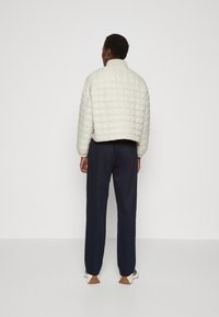 Person med kort hår iført en hvit quiltet jakke, marineblå bukser, hvite sneakers med brune såler, vendt mot en enkel hvit vegg.