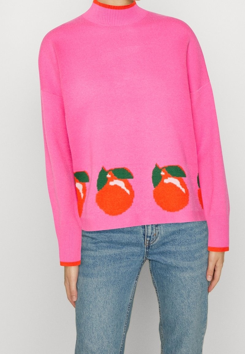 Femme portant un pull rose vif avec des motifs de fruits orange et de feuilles vertes le long de l'ourlet, associé à un jean bleu.