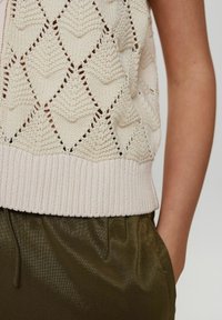 Stickad ärmlös topp i krämfärg med diamantmönster, med en ribbad nederkant. Kombinerad med olivgröna, silkesliknande byxor.