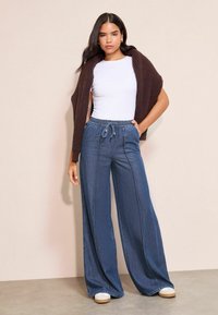 Széles szárú farmernadrág húzózsinóros derékrésszel és kontrasztos varrással, fehér pólóval és barna kötött kardigánnal kombinálva.