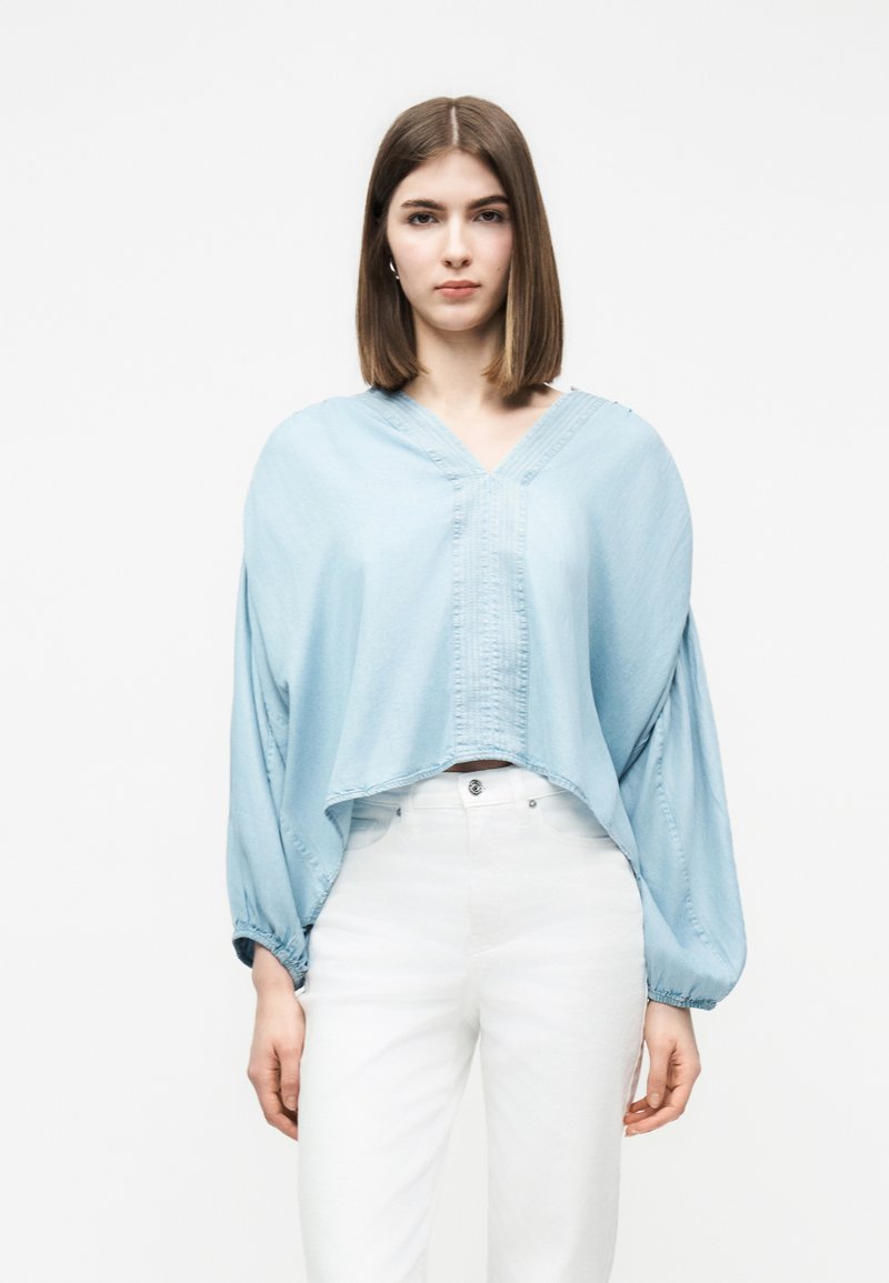 Γυναίκα με ίσια καστανά μαλλιά φοράει ανοιχτό μπλε φαρδιά μπλούζα με V λαιμόκοψη και μπαλόν μανίκια, καθώς και λευκό παντελόνι ψηλόμεσο, μπροστά σε λευκό φόντο.