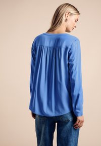 Blå långärmad blus med en mjuk textur, med en plisserad bakdesign och rundad fåll. Kombinerad med ljusblå denimjeans.