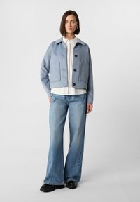 Helles blaues, tailliertes Jacket mit Kragen, schwarzen Knöpfen und zwei großen Fronttaschen. Kombiniert mit weit geschnittenen Denim-Jeans und schwarzem Schuhwerk.