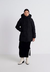 Svart vadderad jacka med huva, snörningar och fickor fram, bärs över en åtsittande svart kjol med en sidoklyfta och beige anklarstövlar.