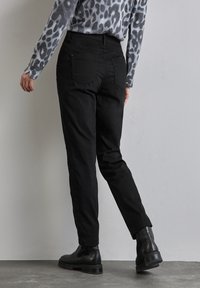 Pantalones negros con un corte entallado, que presentan bolsillos traseros, y combinados con botines negros. El fondo es liso y neutral.
