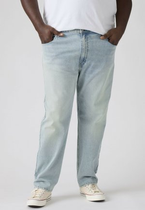 Man draagt lichtblauwe loszittende spijkerbroek en witte sneakers, staat met handen in zakken tegen een effen achtergrond.