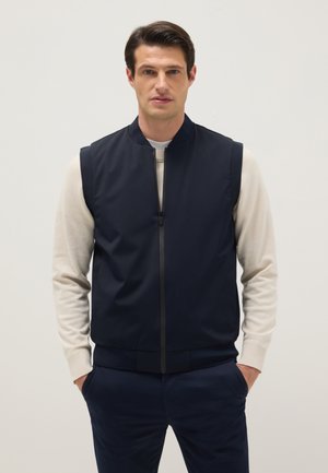 Man wearing navy blue zip-up vest over beige long-sleeve shirt with hands in navy pants pockets against plain background.