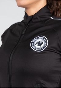 Chaqueta deportiva negra con un alto cuello, que presenta un logotipo blanco circular en el pecho, cierre de cremallera y tejido texturizado. Raya de acento blanca.