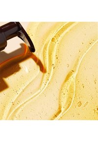 Amberfärgad vätska med en mjuk textur och bubblor, som flödar från en mörk pumpdispenser mot en ljus bakgrund.