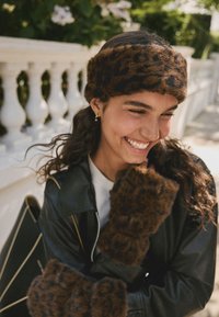 Pannband och fingervantar i brun ton med leopardmönstrad fuskpäls, kombinerade med en svart skinnjacka, mot en suddig utomhusbakgrund.