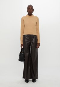 Beige kötött pulóver magas nyakkal, széles lábú barna bőrnadrággal párosítva. A modell egy fekete táskát tart arany részletekkel.