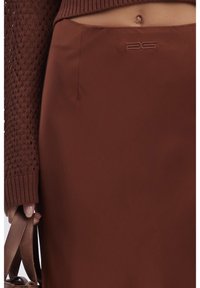 Boven de knie gelegen, bruine rok van een gladde, glanzende stof met een subtiele logo-detail nabij de tailleband. In combinatie met een structureel bruine trui.