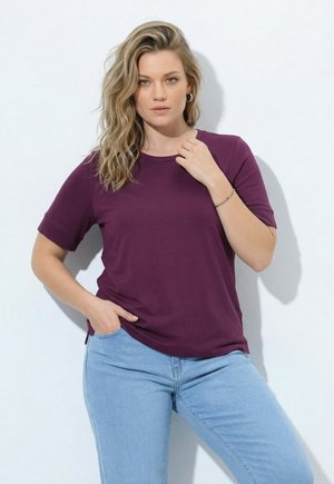Vrouw met golvend blond haar die een paarse korte mouwen shirt en lichtblauwe spijkerbroek draagt, staand tegen een witte achtergrond.