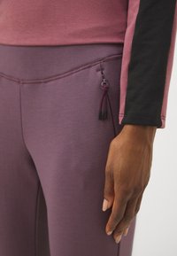 Ljubičaste leggingse za vježbanje izrađene od glatkog materijala, s bočnim džepovima ukrašenim šnurlom i kvačicama, u kombinaciji s ružičastom majicom dugih rukava.