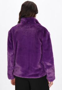 Donna che indossa un giubbotto in pelliccia sintetica viola, con maniche lunghe, vista da dietro su uno sfondo bianco semplice.