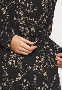 Vestido negro con patrón floral beige. Presenta un diseño envolvente, mangas largas y una cintura fruncida, mostrando una textura de tela suave.