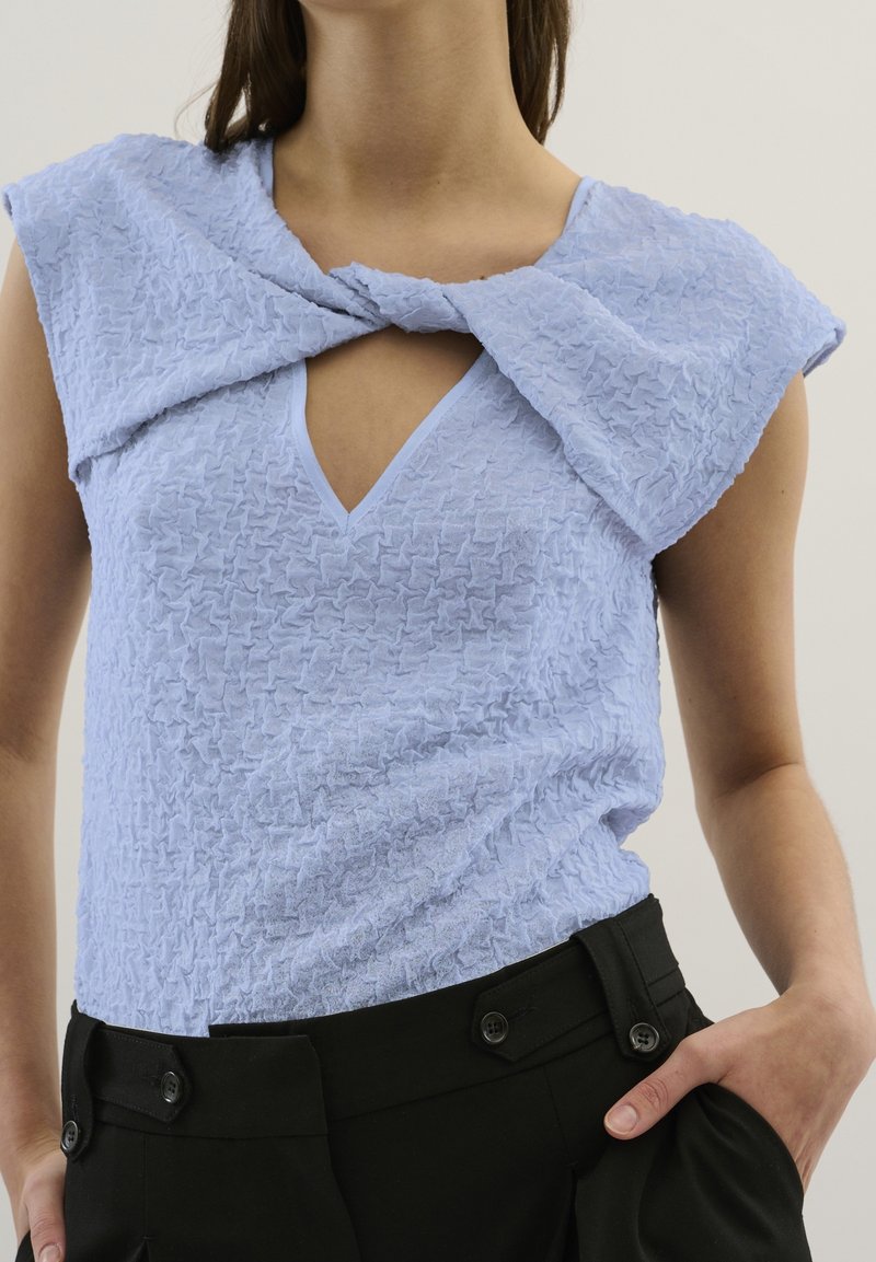 Femme portant un haut sans manches bleu clair texturé avec un détail d'encolure torsadée et un pantalon noir taille haute avec des boutons.