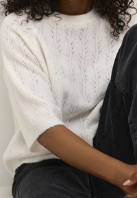 Pull blanc en tricot avec un motif de feuilles texturé, larges manches courtes et ourlet côtelé. Associé à un jean gris foncé.