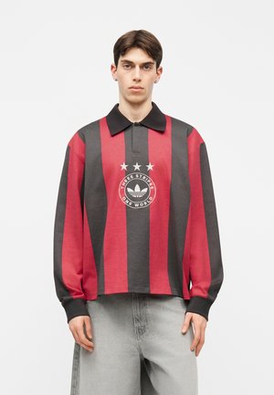 Młody mężczyzna ubrany w czerwoną i czarną koszulkę polo z długim rękawem w pionowe paski z logo „Three Stripes One World” oraz jasnoszare dżinsy.