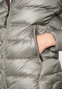 Veste matelassée grise avec fermeture éclair à l'avant, présentant une texture douce et brillante, des coutures horizontales, et une poche latérale avec une fermeture éclair contrastante.