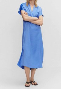 Blaues Midikleid mit kurzen Ärmeln, V-Ausschnitt und seitlichen Schlitzen. Aus glattem Stoff gefertigt, fällt es locker unter die Knie. Kombiniert mit schwarzen Sandalen.