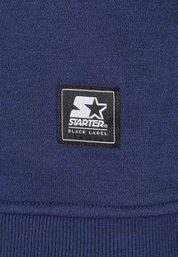 Marineblåt stof med en vævet tekstur, der har et sort rektangulært mærke med teksten "STARTER BLACK LABEL" i hvid skrift.