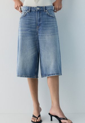 Oseba, oblečena v ohlapne, svetlo modre denim bermuda hlače, z rokami v žepih in črne sandale na peto z vrvico, na enobarvnem ozadju.