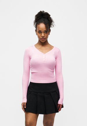 Ung kvinde med krøllet hår iført en tætsiddende lyserød ribbet langærmet top og sort plisseret mininederdel, stående mod en hvid baggrund.