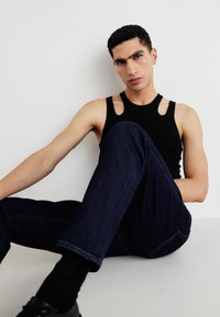 Čierny bezrukávový top s výrezmi na pleciach, skombinovaný s tmavomodrými džínami. Model sediaci na hladkom povrchu. Minimalistický dizajn.