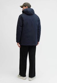Marinefarbener gepolsterter Mantel mit Kapuze, ausgestattet mit einer glatten Textur und lockerer Passform, kombiniert mit weiten schwarzen Hosen und hellen Schuhen.