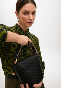 Svart vävd läderväska med guldkedja och flätat handtag. Modellen bär en grön leopardmönstrad skjorta med svarta knappar.