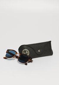 Ochelari de soare Ray-Ban cu rame din fâșie țestoasă, lentile întunecate și accente aurii, plasați lângă un etui din piele neagră cu un logo circular.
