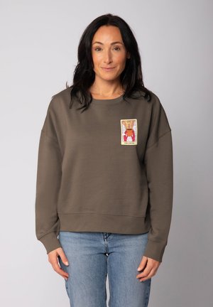 Woman with dark hair wearing a brown sweatshirt with a colorful coffee-themed patch on the chest, paired with light blue jeans.