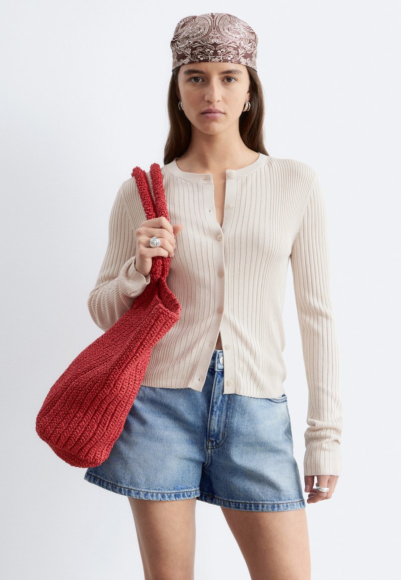 Vrouw met een patroon hoofddoek, beige geribde cardigan, denim shorts, die een grote rode gebreide tas over haar schouder draagt.