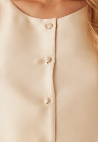 Blusa beige con una texture liscia, caratterizzata da un colletto rotondo e da tre bottoni decorativi sulla parte anteriore.