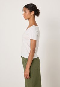 Bijela majica kratkih rukava s širokim izrezom, izrađena od mekanog materijala. Nošena s hlačama opuštenog kroja maslinasto zelene boje koje imaju bočne džepove.