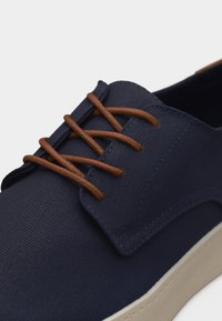 Chaussure décontractée bleu marine en tissu texturé, avec des lacets marron et une semelle en caoutchouc blanche, présentée en gros plan sur un fond blanc.
