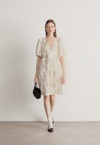 Μπεζ φόρεμα με βαθύ V-λαιμόκοψη, φουσκωτά μανίκια, υφή δαντέλας και λαμπερά στοιχεία, συνδυασμένο με μαύρη τσάντα και λευκές κάλτσες.