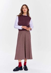 Slivkový, chĺpkový sveter bez rukávov prenesený cez bielu košeľu s golierom, v páre s dlhou, vzorovanou sukňou v červenej a čiernej farbe. Čierne platformové topánky a červené ponožky.