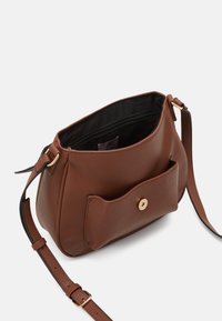 Sac en bandoulière en cuir marron avec une finition texturée, de forme arrondie, une poche avant avec fermeture à pression, et une doublure intérieure noire.