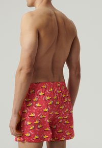 Męski tors widoczny z tyłu, ubrany w czerwone kąpielówki w żółte kaczuszki z napisem "LATO" na jednolitym tle.