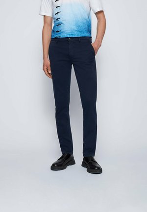 Homme portant un pantalon slim bleu marine, des chaussures habillées noires et un t-shirt dégradé blanc et bleu avec des motifs de poissons, debout avec une main dans la poche.
