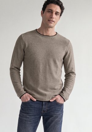 Mann trägt einen beigen Langarmpullover und blaue Jeans, die Hände in den Taschen, steht vor einem schlichten hellen Hintergrund.