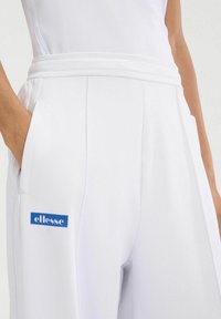 Pantalones blancos con una textura suave, que cuentan con un logo de ellesse azul en el bolsillo izquierdo y una cinturilla estructurada para un ajuste cómodo.